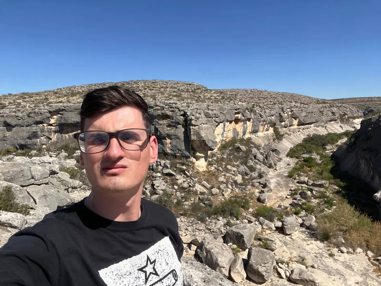 A low-angle self-portrait of a man with glasses against a backdrop of steep, rugged, multi-colored canyon walls under a clear blue sky.