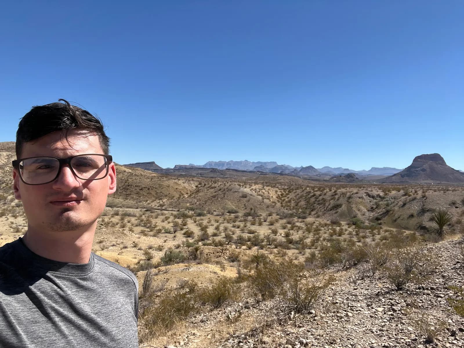 A man stands on a rocky desert path with sprawling shrubs and mountains in the distance under a bright sun.