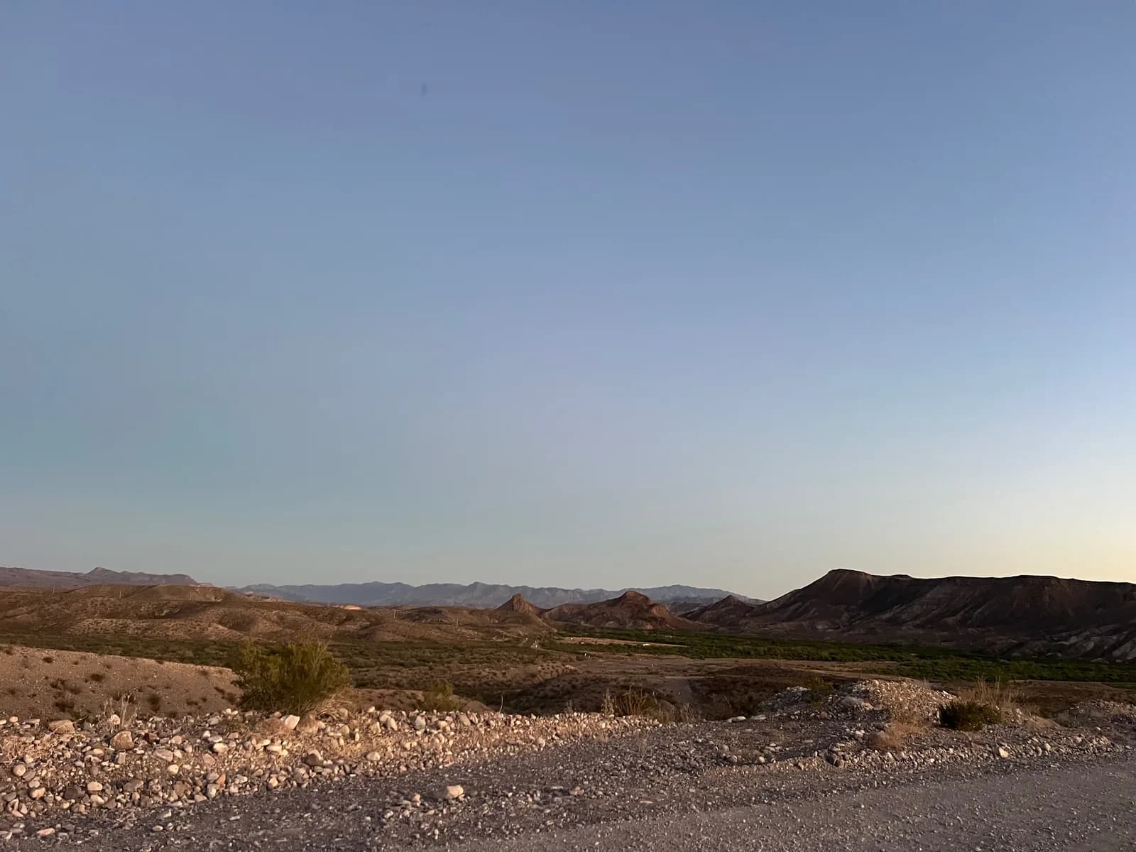 A vast desert landscape under a pale evening sky, showing rolling brown hills, a gravel roadside in the foreground, and distant mountains.