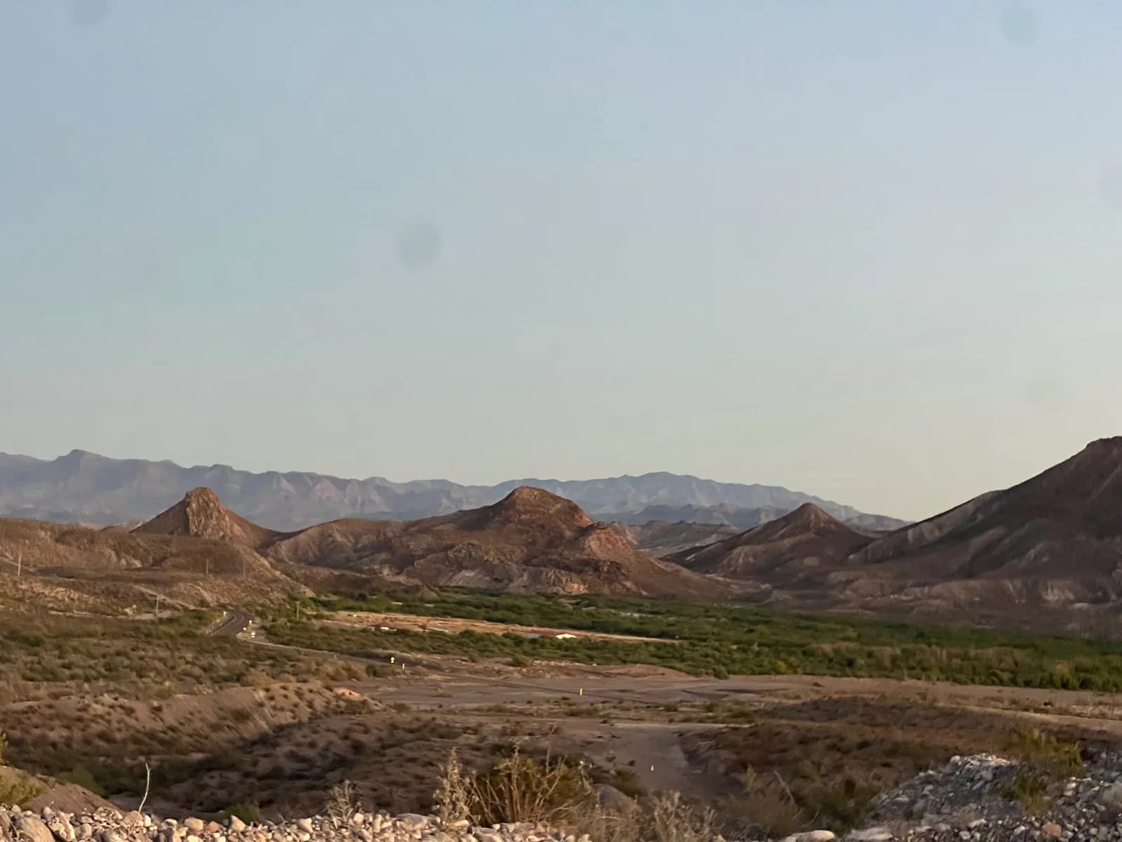 A wide desert landscape at dusk showing rolling brown hills and distant hazy blue mountain ranges under a pale evening sky.