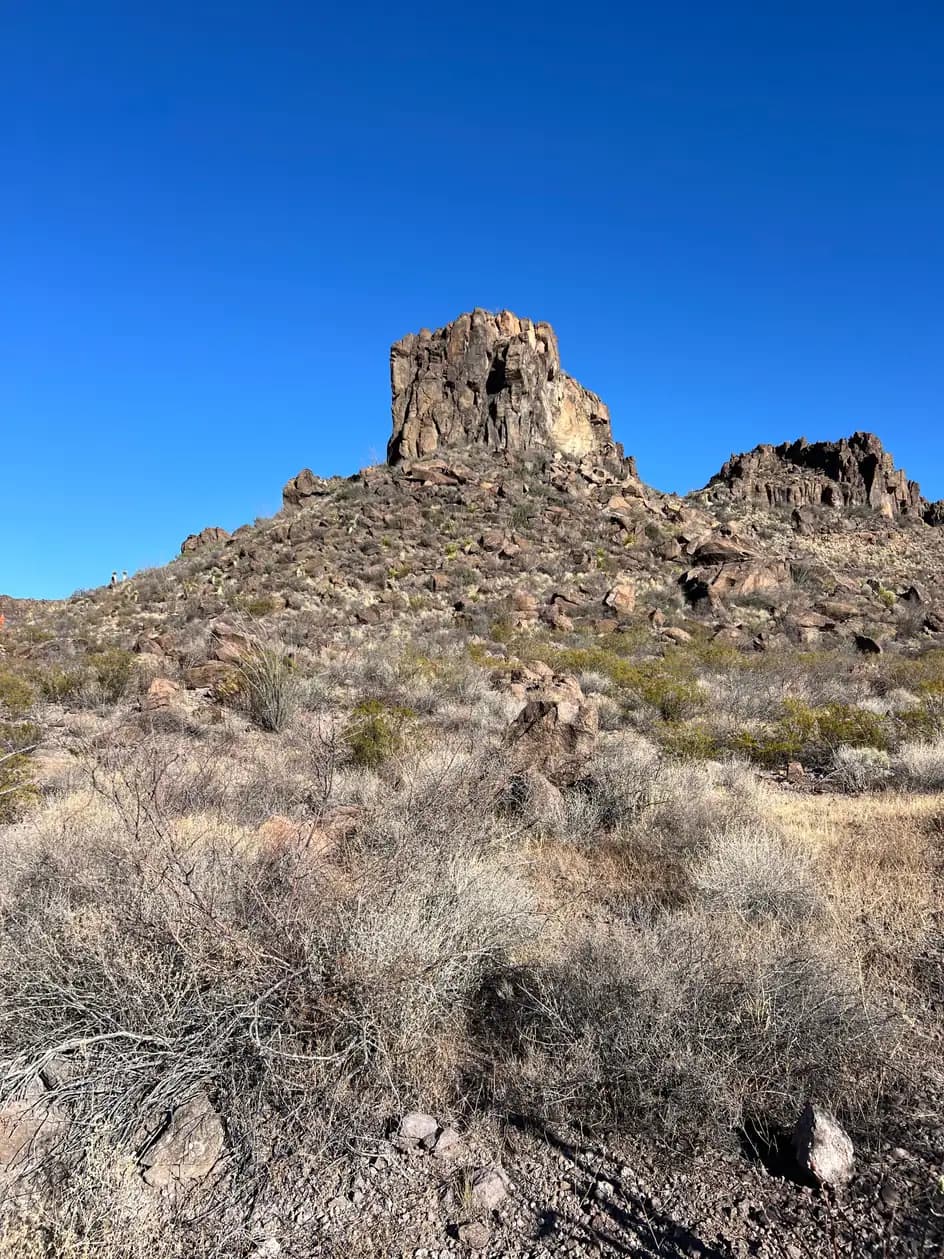 A towering, craggy rock peak rises above a desert slope covered in dry brush, cacti, and loose stones against a clear blue sky.