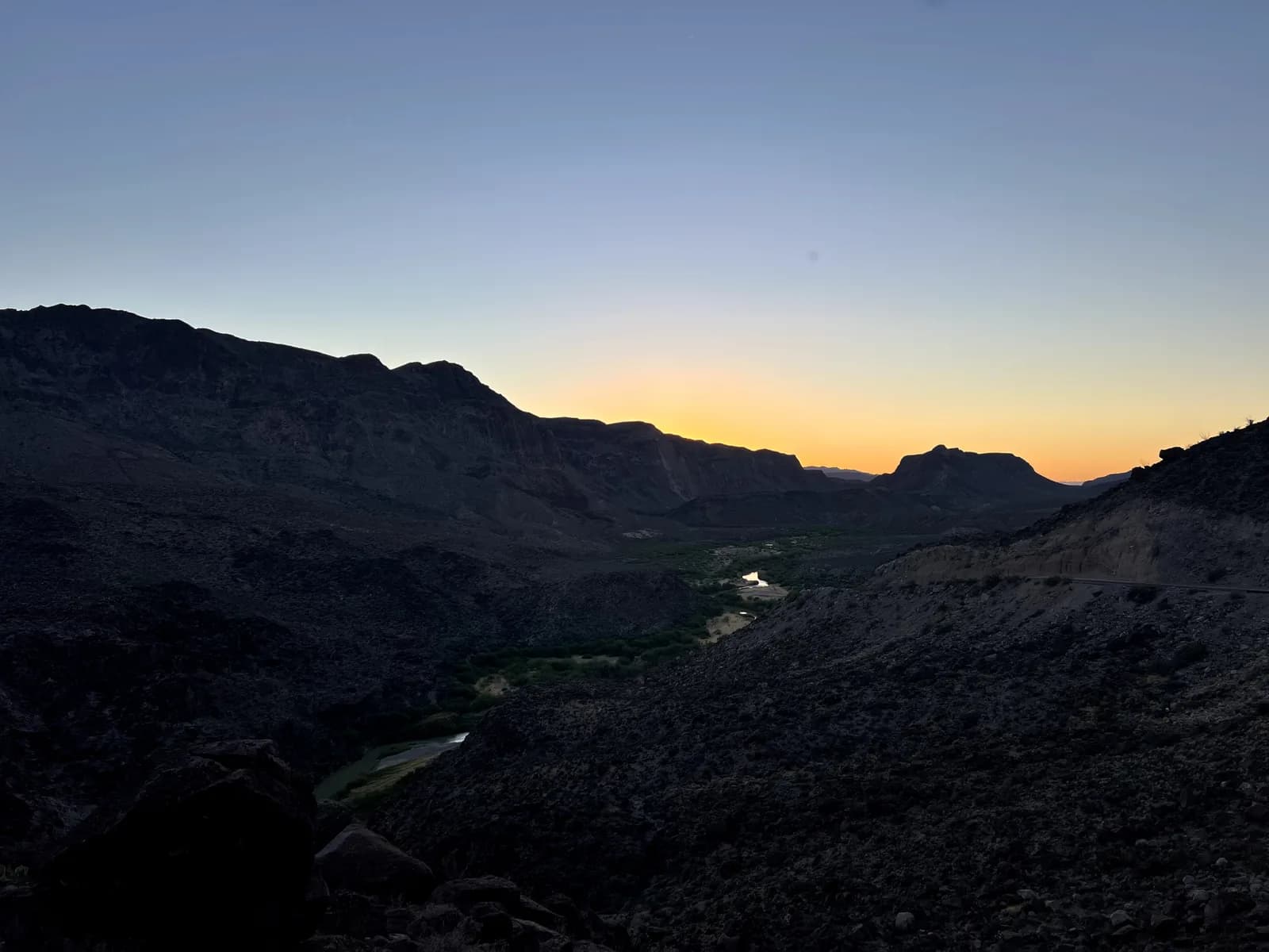 A sunset view from a high desert overlook, showing a glowing orange horizon behind dark mountain silhouettes and a winding river in the valley below.