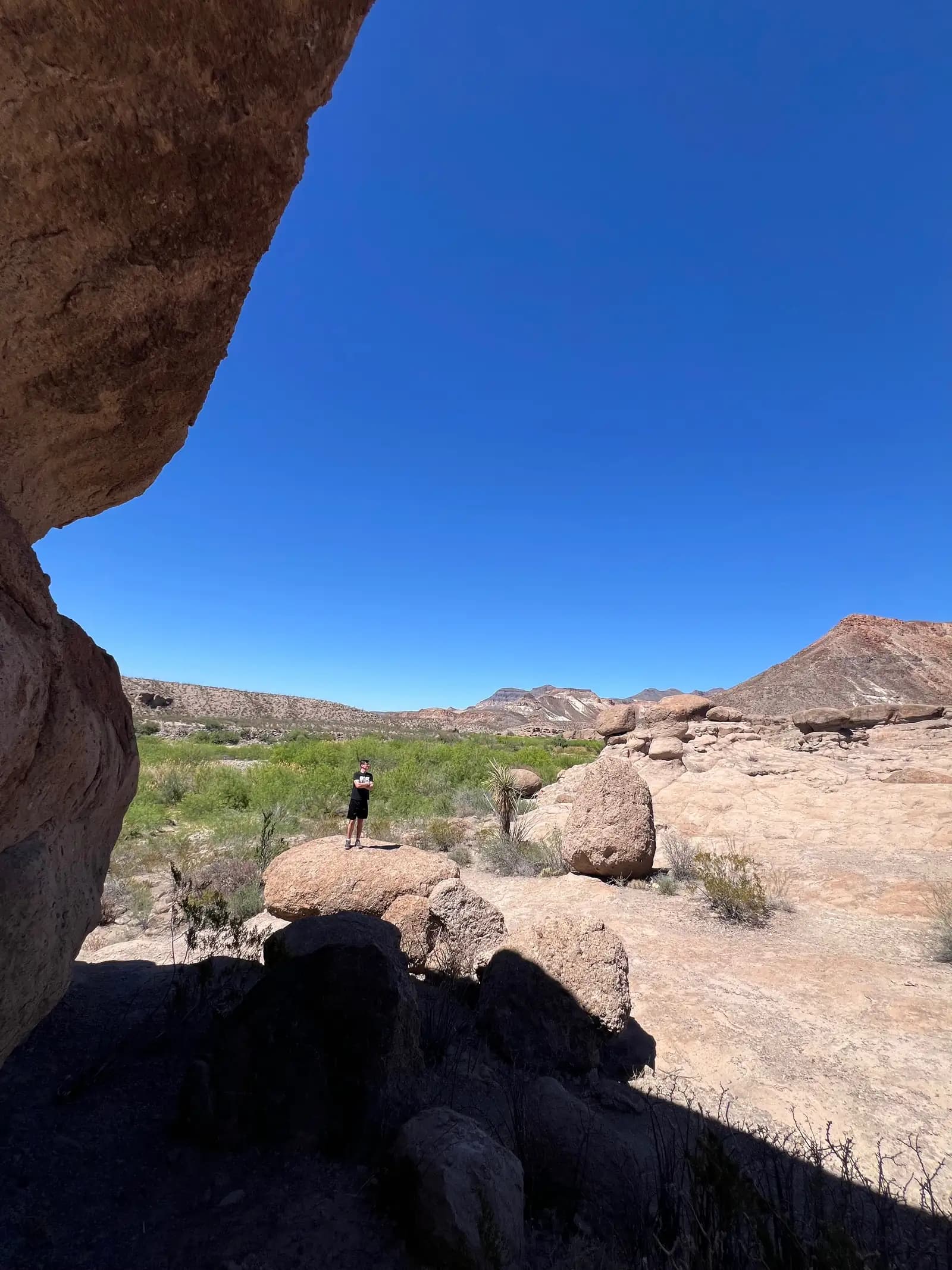 A view from under a rock overhang looking out toward a desert valley filled with unique rock formations, green shrubbery, and distant mountains under a clear blue sky.