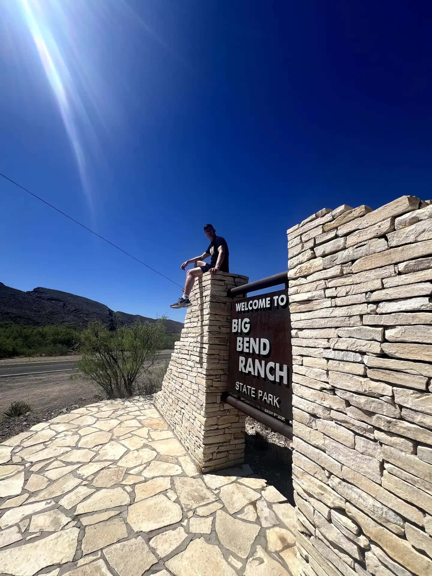 A man sits atop a stone pillar at the entrance of Big Bend Ranch State Park under a clear blue sky, next to a sign that reads "Welcome to Big Bend Ranch.