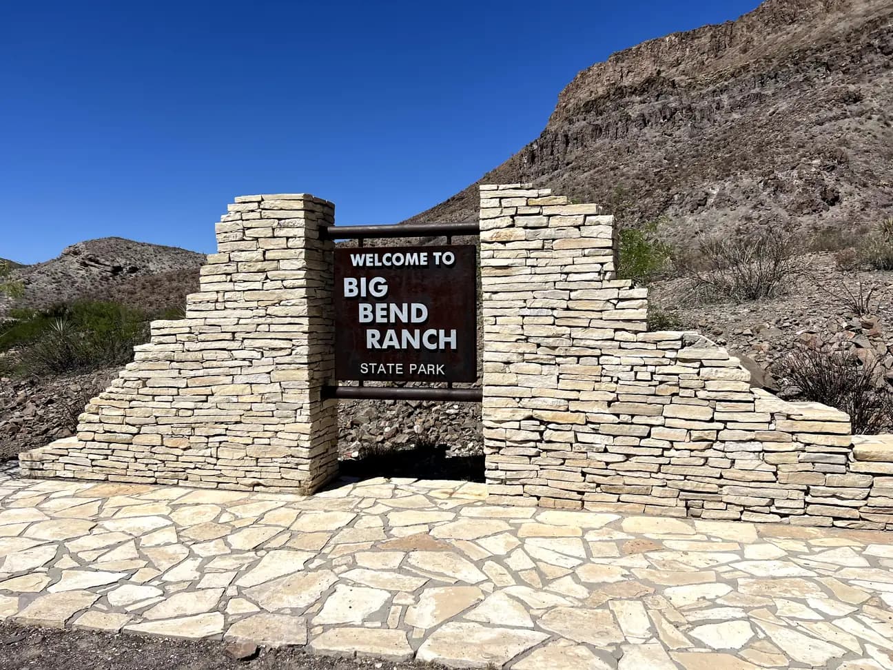 A weathered, stacked-stone entrance gate holding a metal sign that says "WELCOME TO BIG BEND RANCH STATE PARK," set against a desert mountain backdrop.
